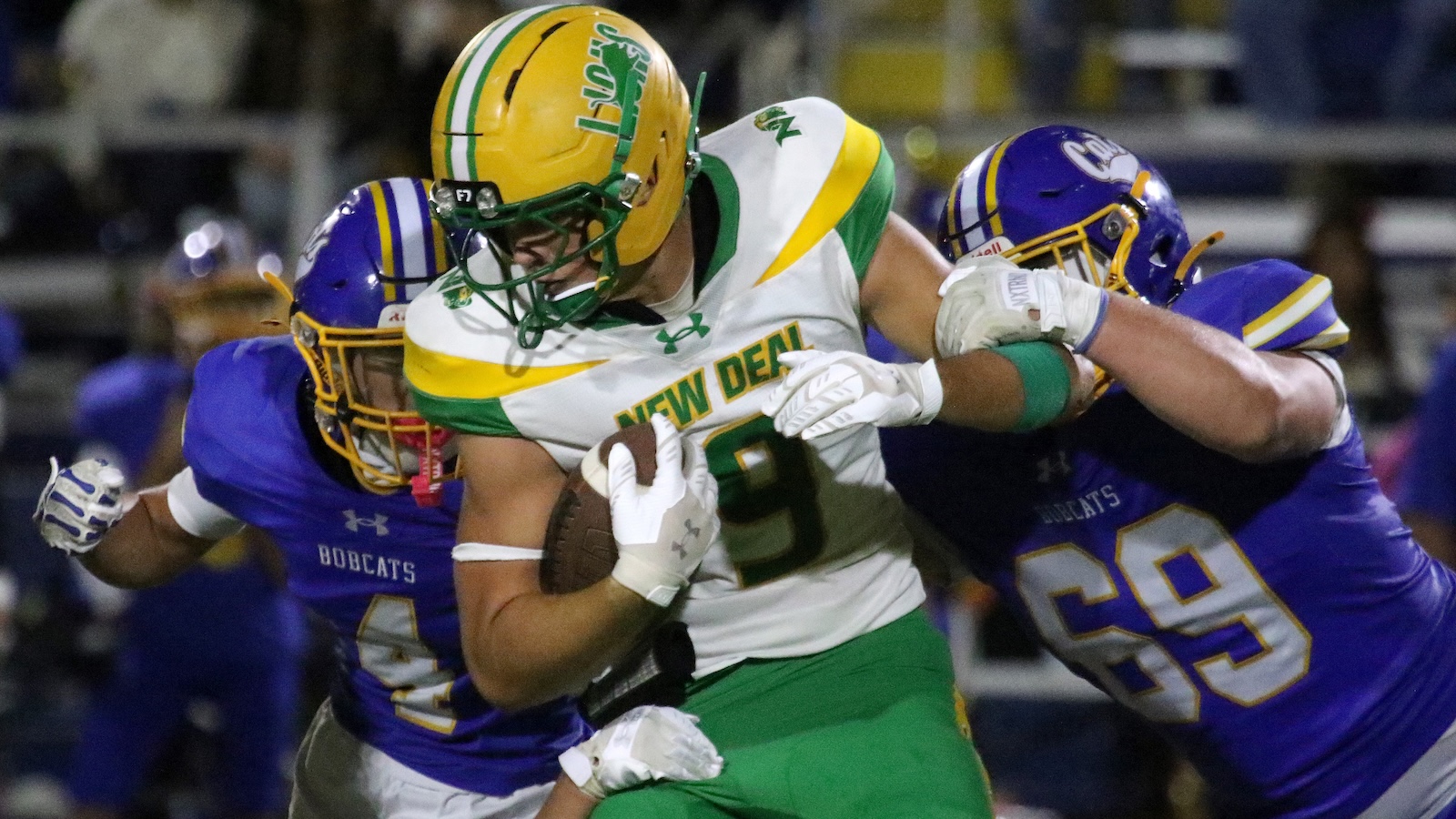 Football player in white-and-green jersey carrying the ball as two purple-clad defenders tackle him.