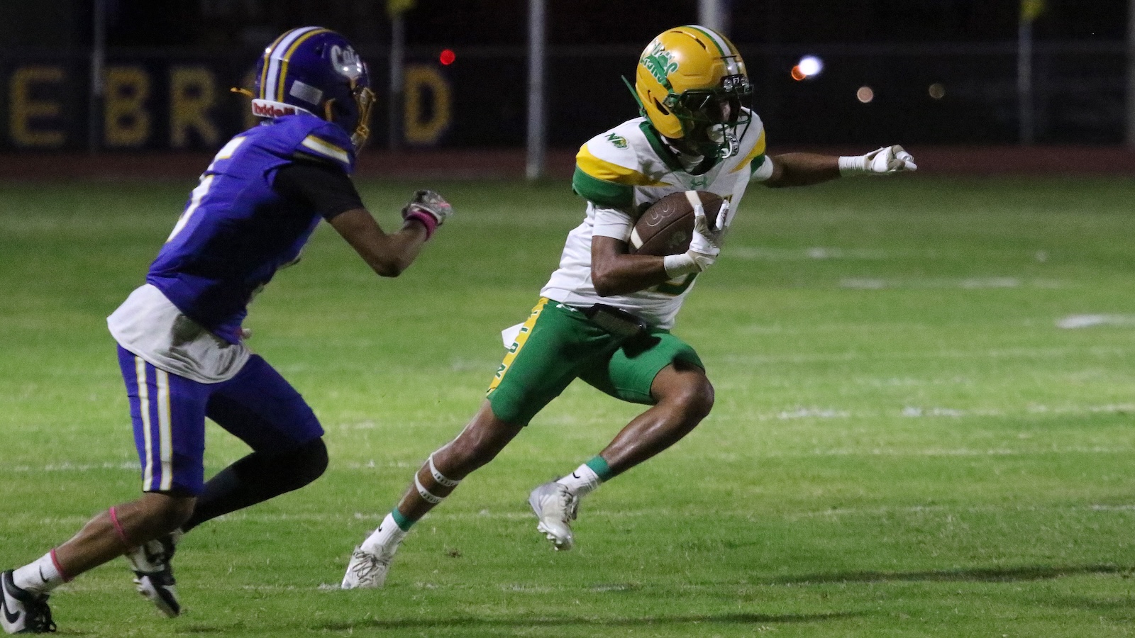 Football player in white and green runs with the ball, dodging a defender in purple at night on a grassy field.