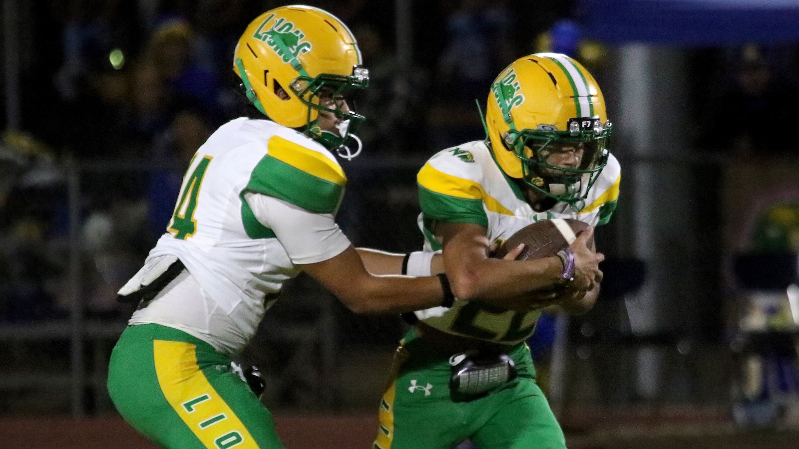 Two football players in green and yellow uniforms compete for the ball; one tackles while the other carries the football through a game at night