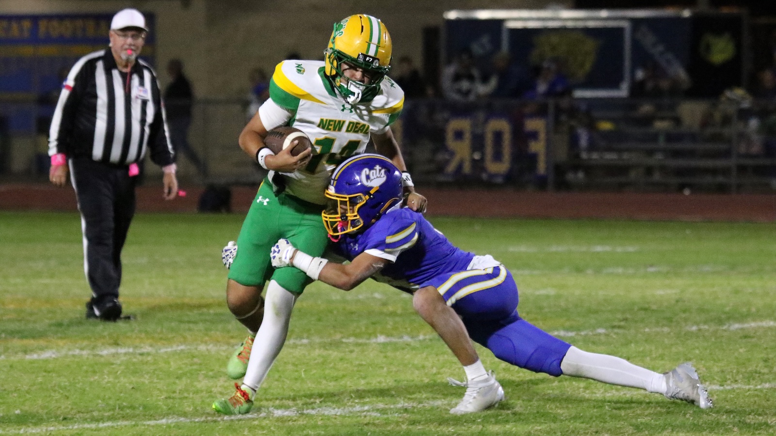 Football player in green and white (New Deal) runs with the ball as a purple-clad defender dives in for a tackle at night on a grassy field