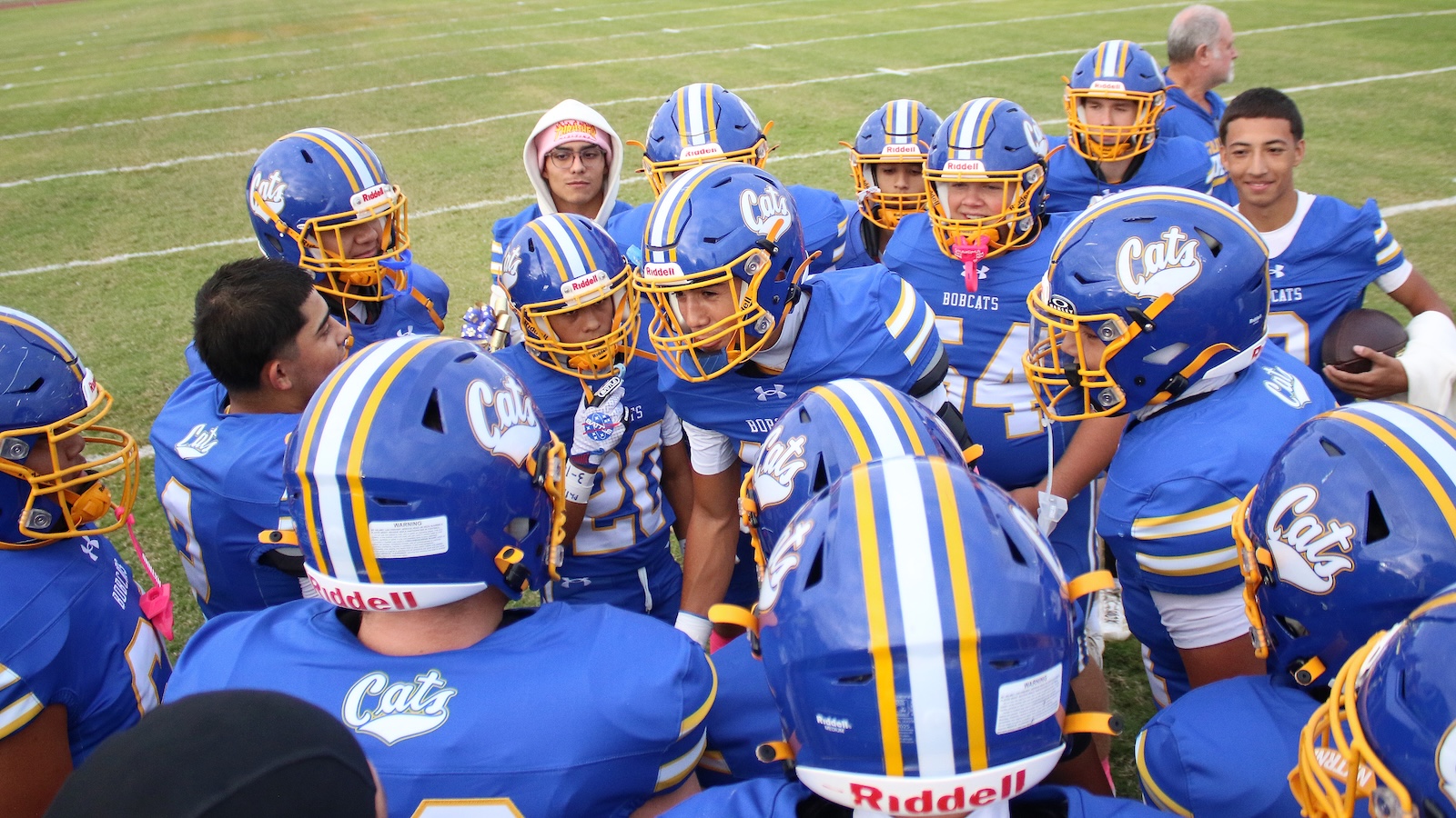 Blue and gold football players huddle on a grassy field before the game, forming a tight circle.