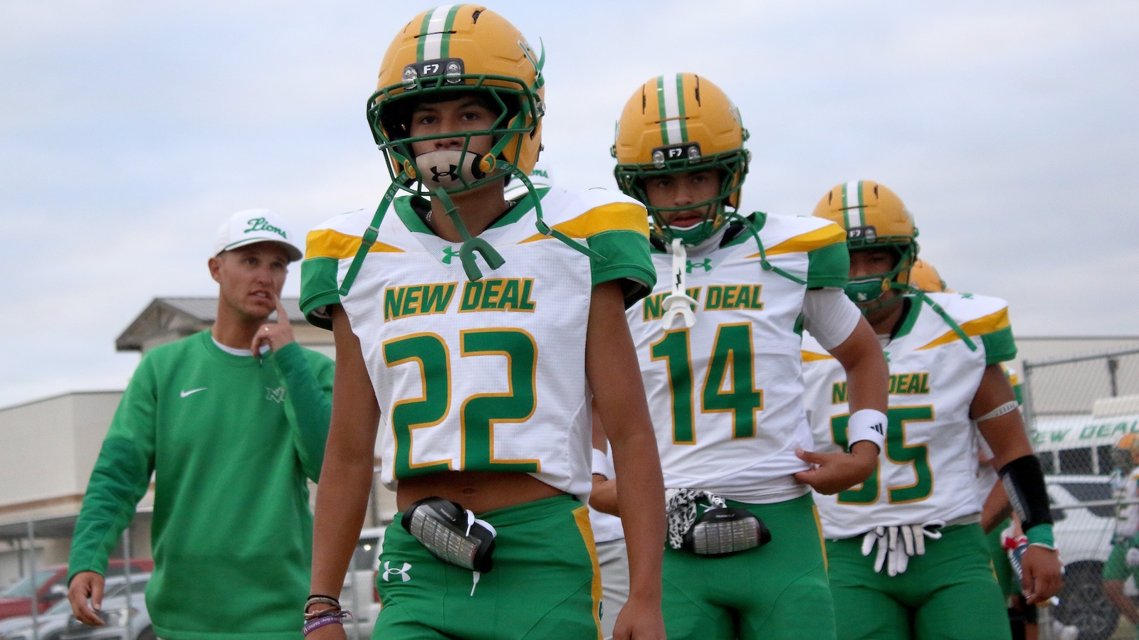 Young football players in green and white uniforms walk onto the field; front player wearing #22, yellow helmet with green stripe. Behind him are teammates with #14 and #95.