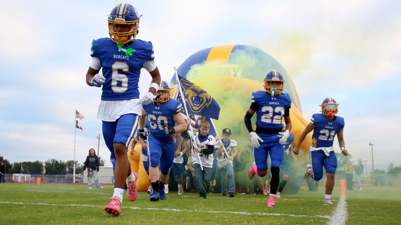 Youth football players in blue uniforms sprint onto the field through a smoke-filled inflatable tunnel at game time.