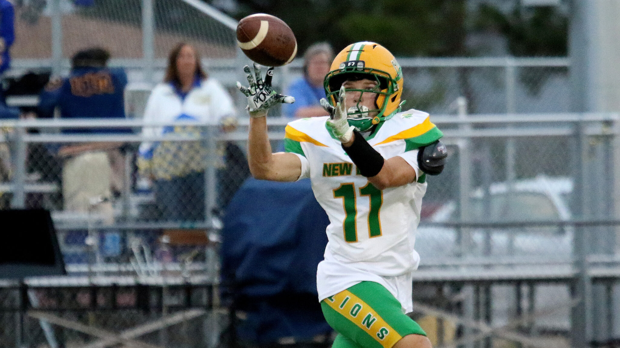 100326 - New Deal vs Sunray - Ethan Mahan | Press Pass Sports American football player in a white uniform with green and yellow accents reaches to catch a pass as the ball descends toward him.