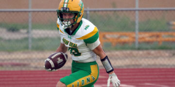 Football player in green and gold uniform runs with the ball on a track field ground surface in action.