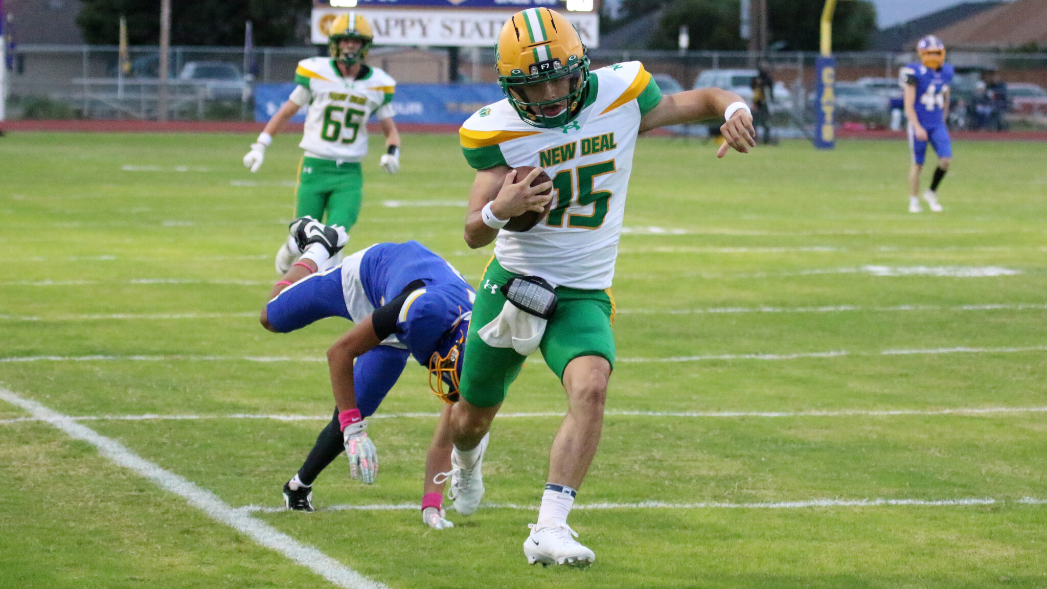 100326 - New Deal vs Sunray - Jackson Attebury | Press Pass Sports Football player in white and green (number 15) runs with the ball, sprinting down the field as a blue-clad defender falls behind on a grass field.