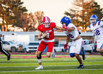 0R8A8541 | Press Pass Sports Football game: a red-clad player runs with the ball pursued by white-uniform teammates on a green turf field under a sunny sky.