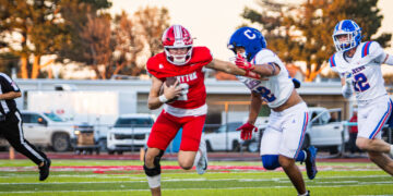 Football game: a red-clad player runs with the ball pursued by white-uniform teammates on a green turf field under a sunny sky.