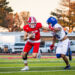 0R8A8541 | Press Pass Sports Football game: a red-clad player runs with the ball pursued by white-uniform teammates on a green turf field under a sunny sky.