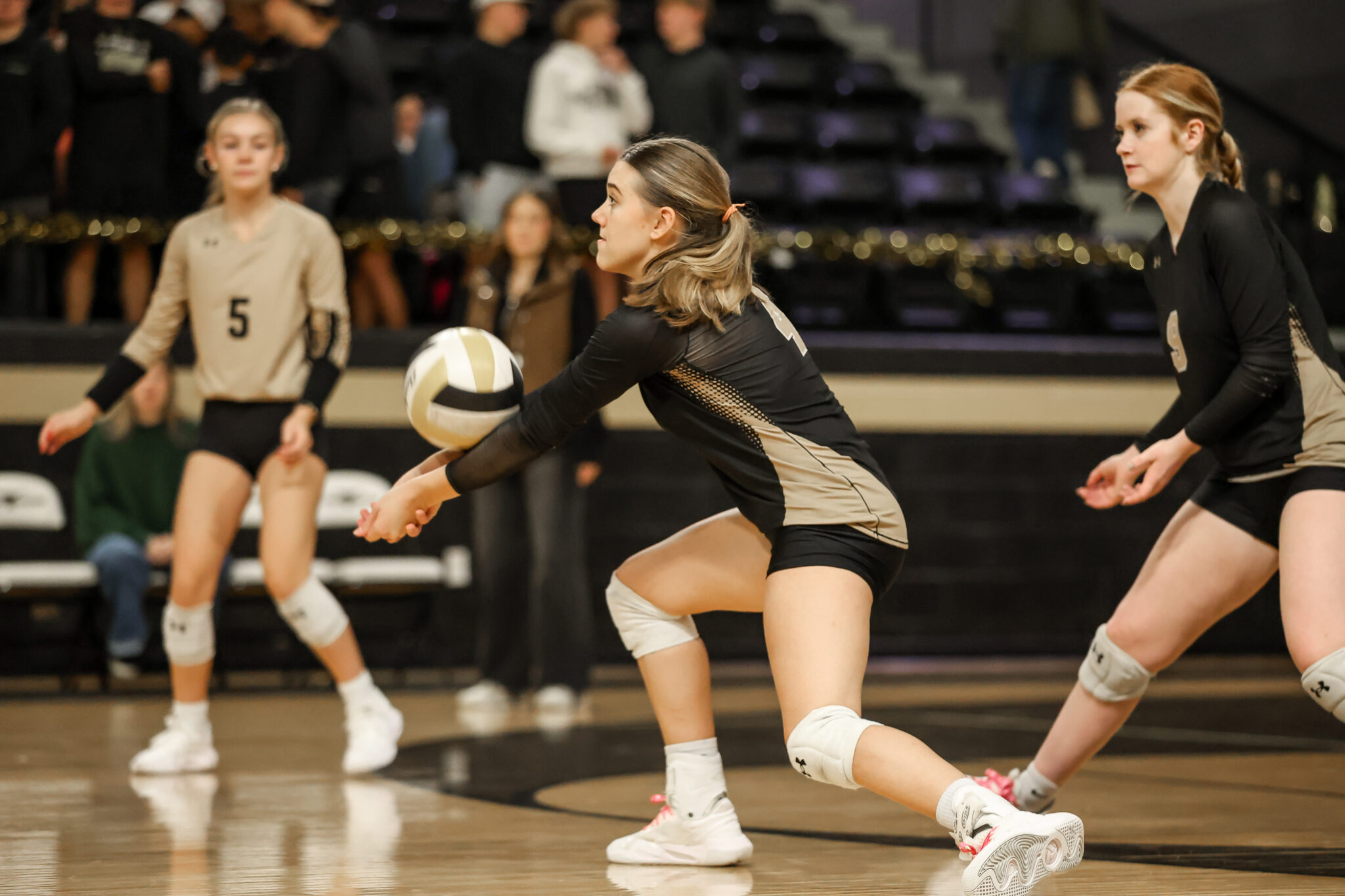 3Q6A3575 | Press Pass Sports Female volleyball players in beige and black uniforms sprint toward the ball on an indoor court as the player in front prepares to pass.
