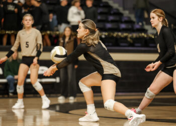 3Q6A3575 | Press Pass Sports Female volleyball players in beige and black uniforms sprint toward the ball on an indoor court as the player in front prepares to pass.