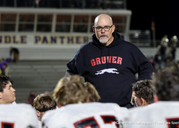 20261002-Gruver-Panhandle-FB-040 | Press Pass Sports Bald man with glasses and gray beard in a dark hoodie with a white horse logo, addressing football players in a stadium.
