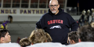 Bald man with glasses and gray beard in a dark hoodie with a white horse logo, addressing football players in a stadium.