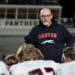 20261002-Gruver-Panhandle-FB-040 | Press Pass Sports Bald man with glasses and gray beard in a dark hoodie with a white horse logo, addressing football players in a stadium.