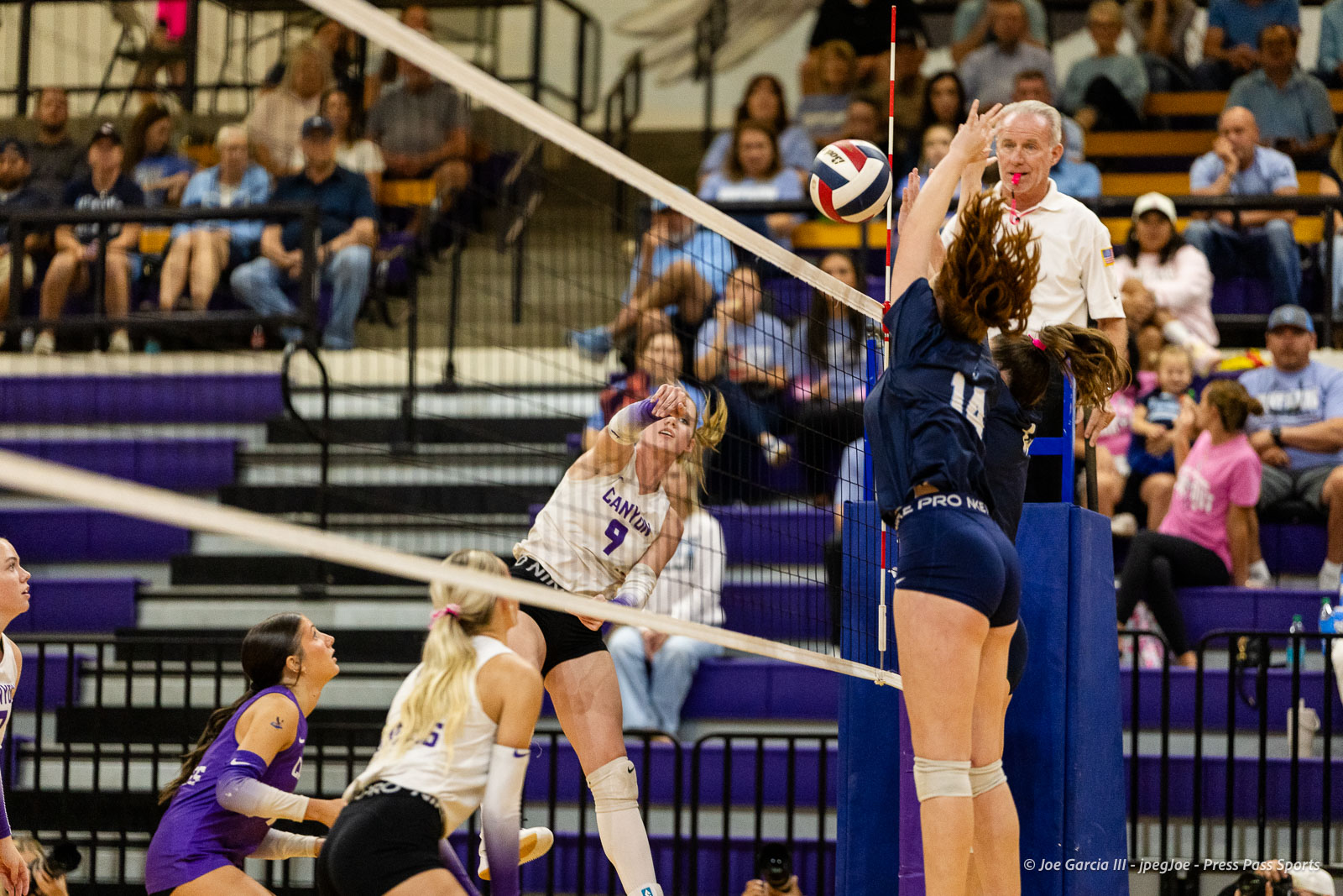 Volleyball action at the net: a white-uniformed player (#9) spikes the ball while a blue-uniformed blocker (#14) jumps to block; referee watches nearby.