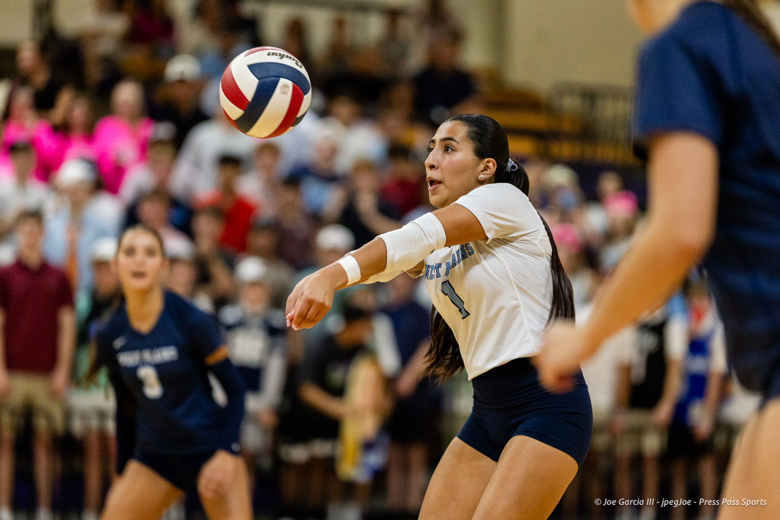 Female volleyball player in a white jersey reaches for the ball during a match, with a crowded gym in the background.