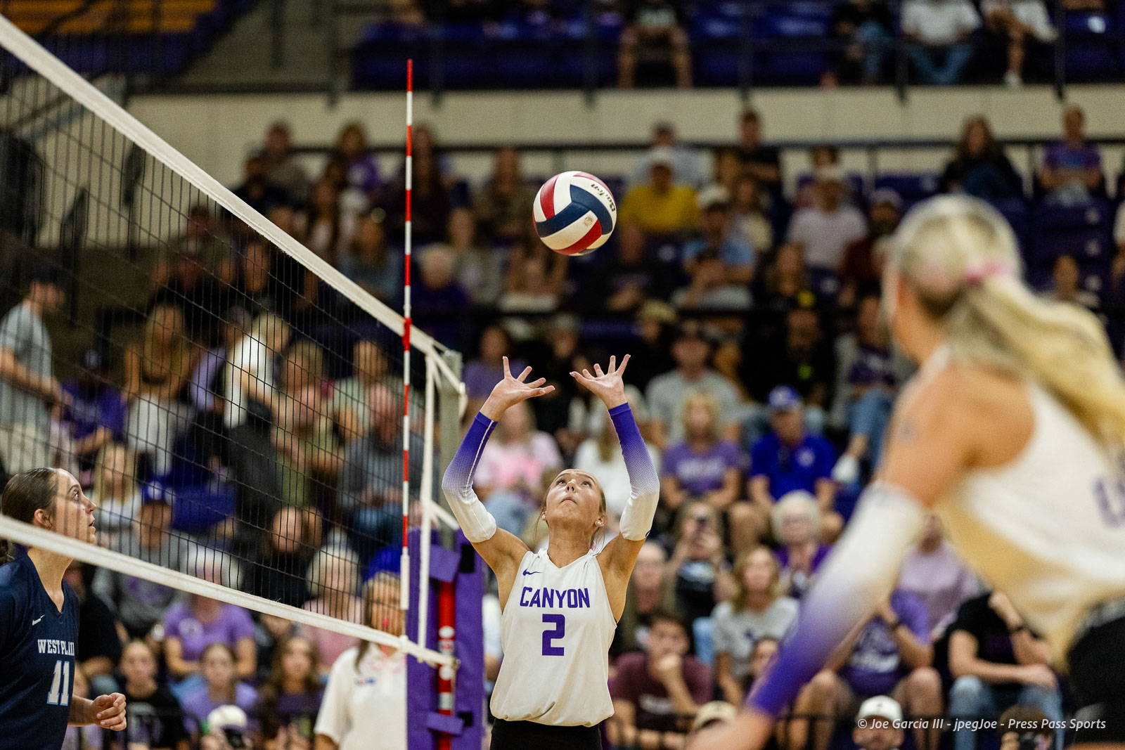 Volleyball player in a white Canyon jersey with number 2 jumps to set the ball over the net as spectators watch.