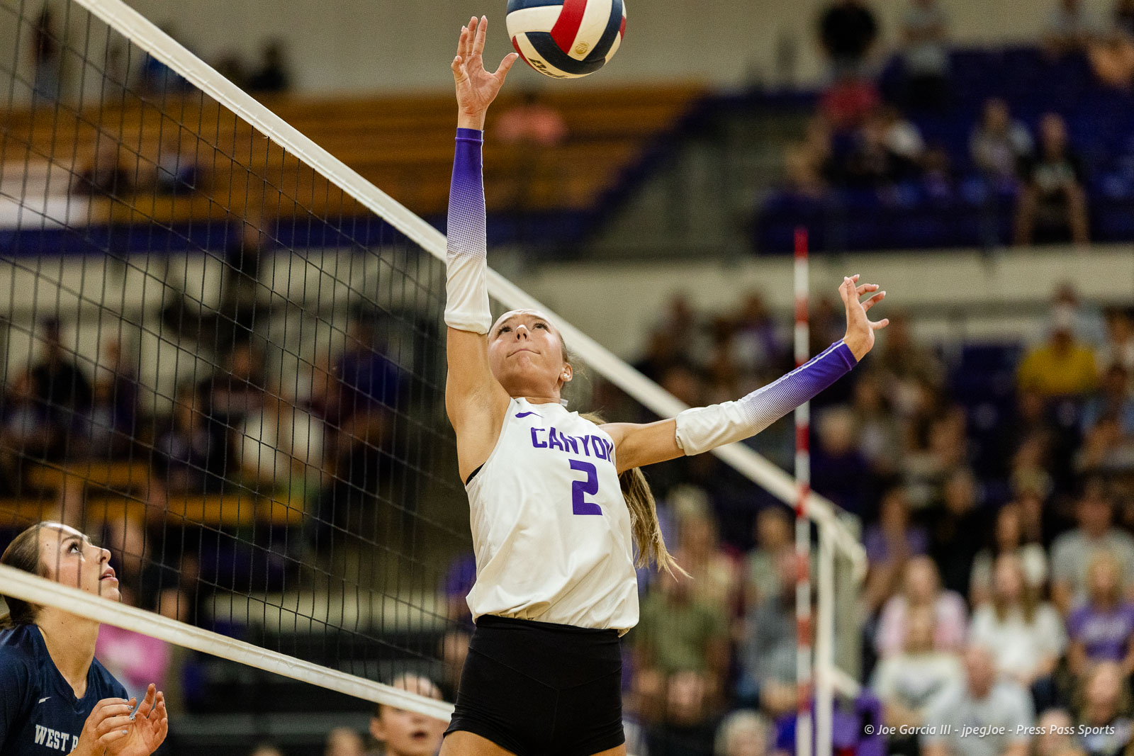 Female volleyball player in white jersey with number 2 jumps to spike the ball over the net, purple sleeves visible, gymnasium crowd in background.