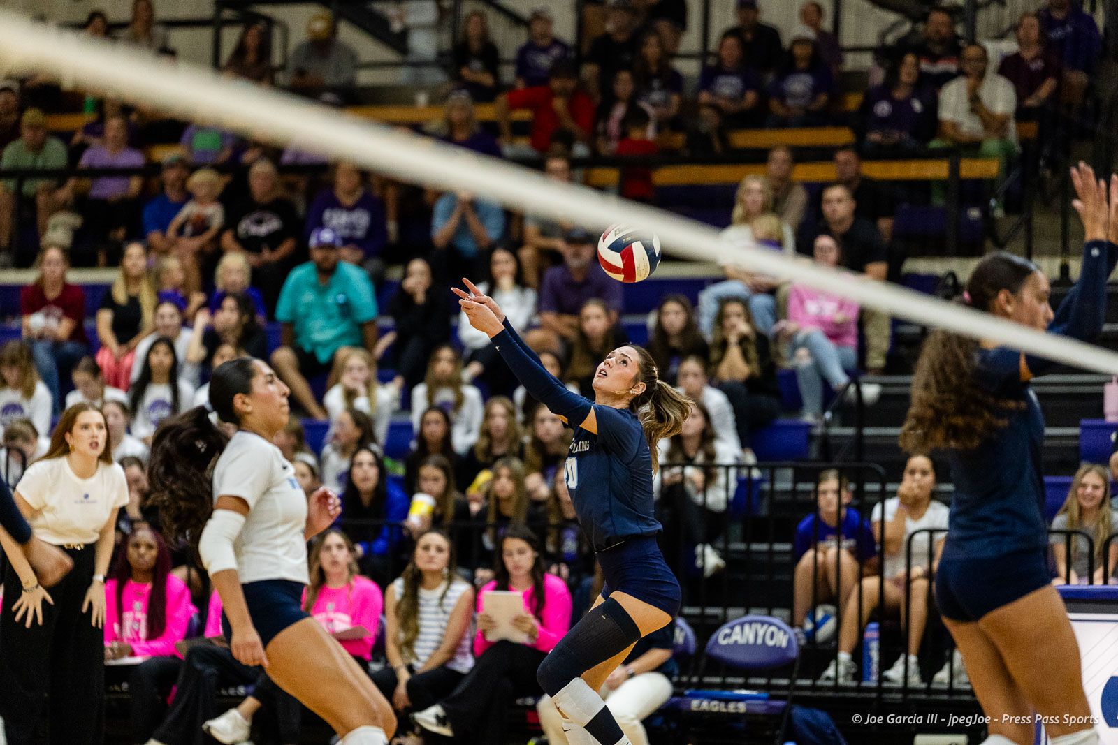 Female volleyball player in navy uniform leaps to strike the ball midair as spectators watch from the bleachers nearby.