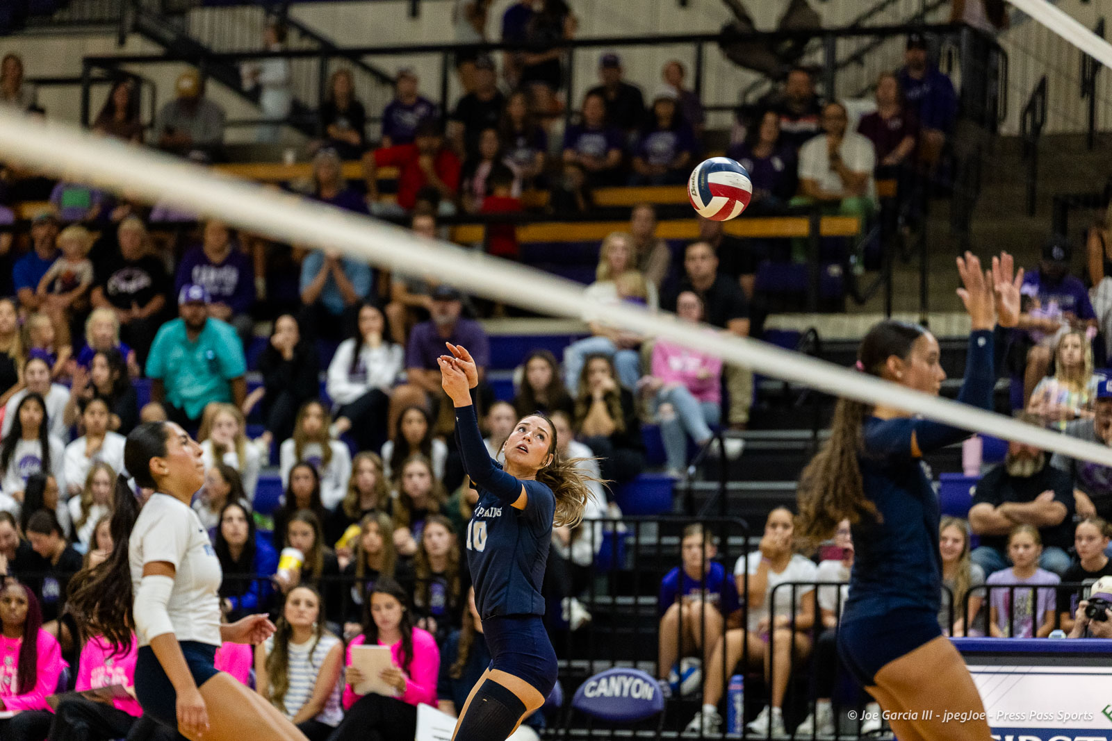 Volleyball player in navy uniform reaching for the ball near the net as the crowd watches from the bleachers below.