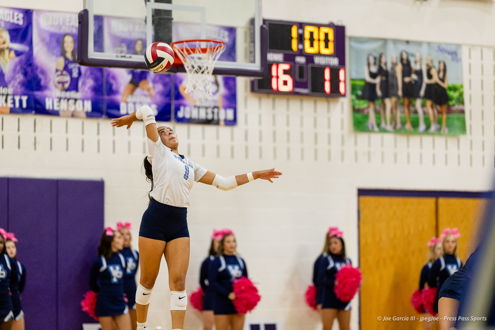 Female volleyball player in a white jersey (#1) jumps to spike the ball during a match in a gym, with banners and cheerleaders in the background.