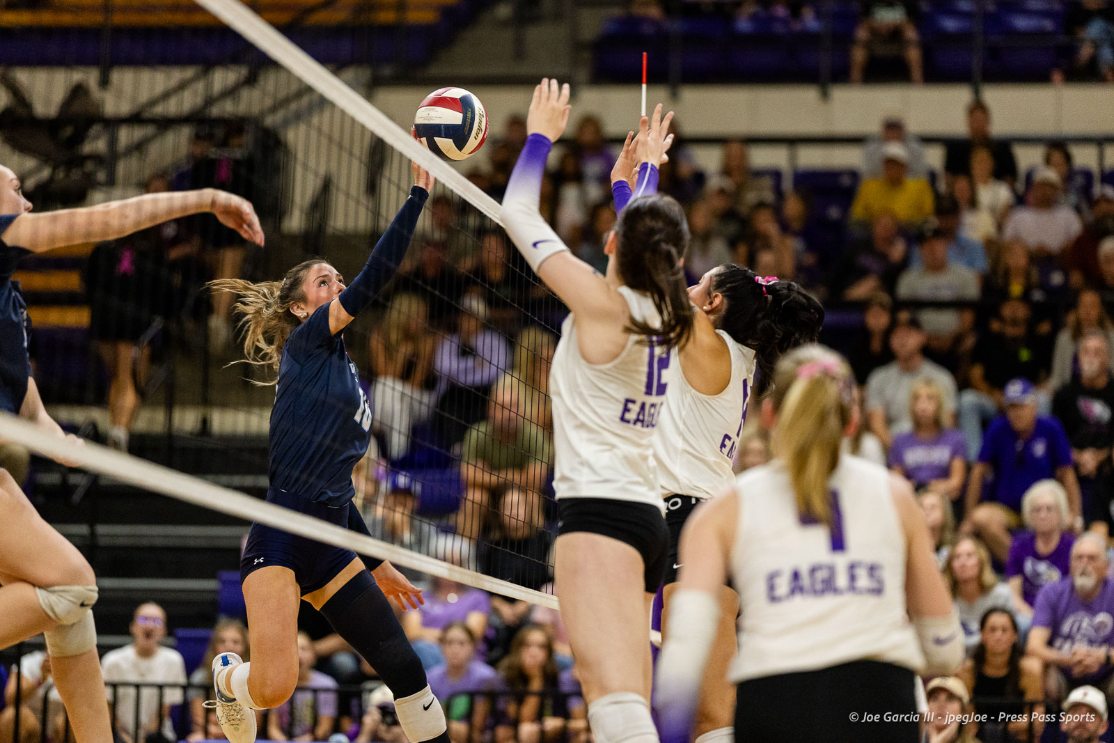 Female volleyball player in navy uniform leaps to spike the ball over the net as white-uniform blockers jump to block, crowd watching in background.