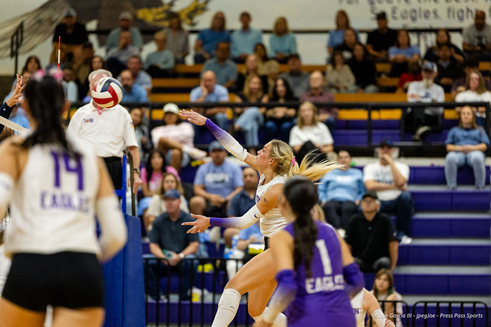 Female volleyball player in white reaching to hit the ball near the net as teammates in purple defend, with a crowded gym audience in the background.