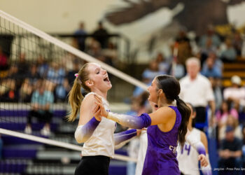 west-plains-vs-canyon-volleyball-jpegJoe-19 | Press Pass Sports Two female volleyball players celebrate with a high-five at the net during a match, one in white and one in purple, with a cheering crowd in the background.