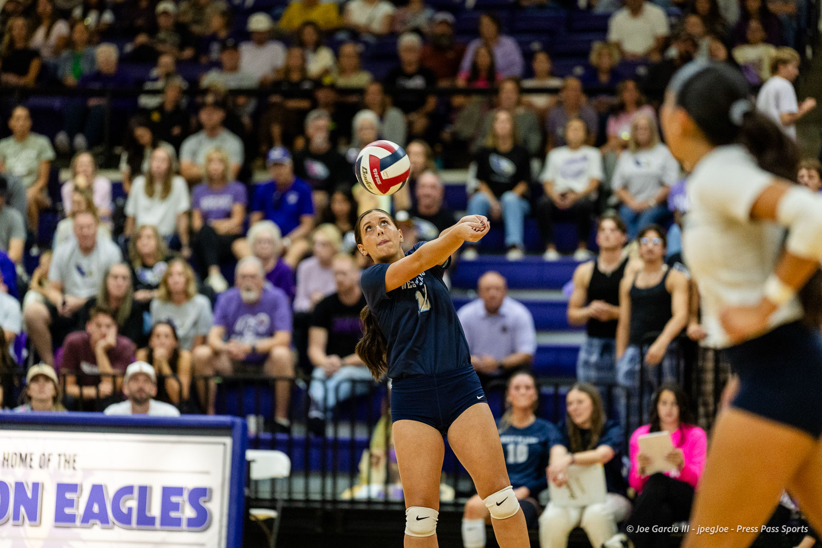 Female volleyball player in navy uniform serves the ball during a match with a crowded gym in the background.