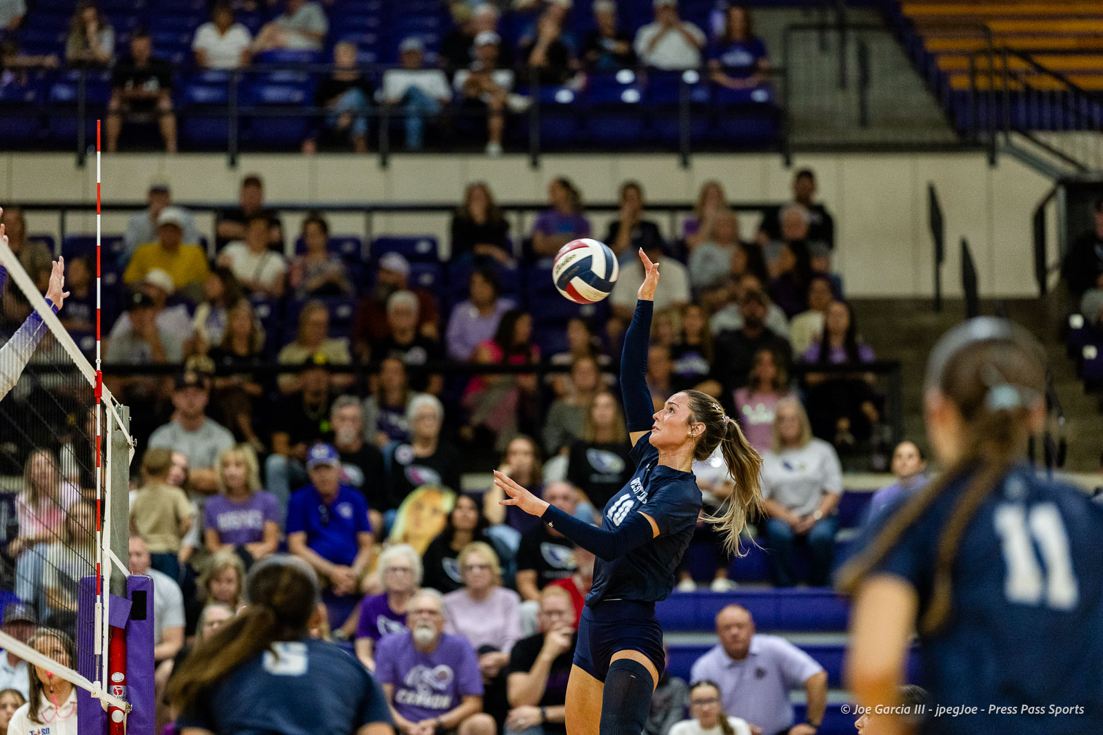 Female volleyball player in navy uniform spikes the ball over the net during a match, crowded gym in the background.