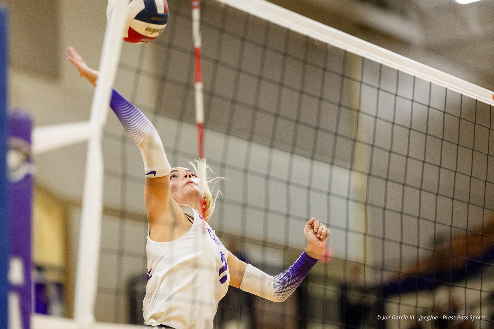 Female volleyball player in a white uniform jumps to spike the ball over the net in an indoor gym.