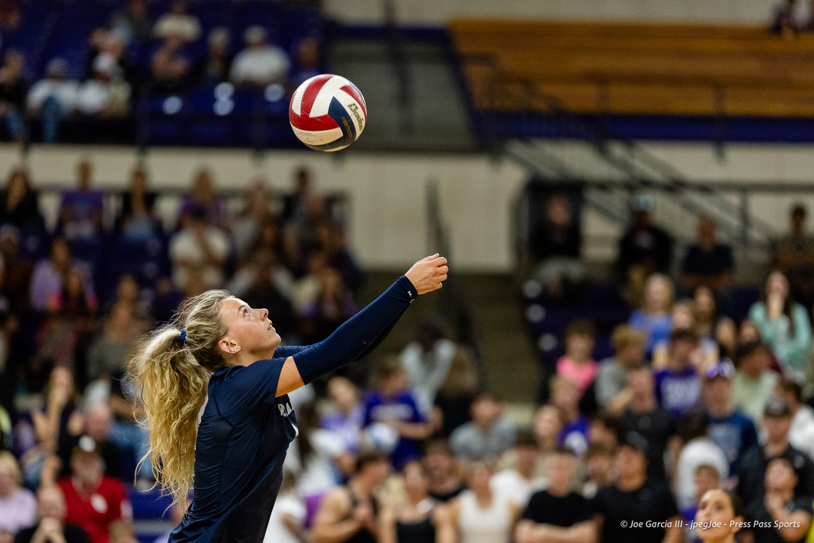 Volleyball player in navy uniform serves the ball in an indoor arena with a cheering crowd in the background, arm extended upward toward the ball.