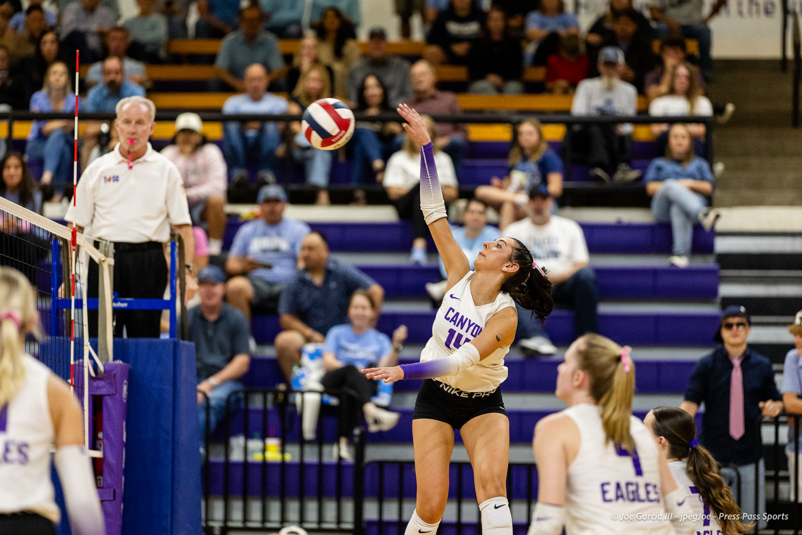 Female volleyball player in a white Canyon jersey leaps to spike the ball over the net as a referee watches, with spectators in the bleachers behind.