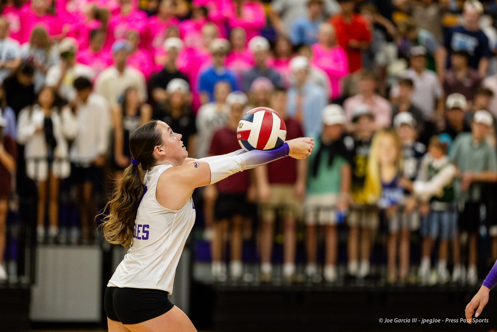 Female volleyball player in a white jersey and black shorts extends to set a red, white, and blue ball as a crowded gym watches from the stands.