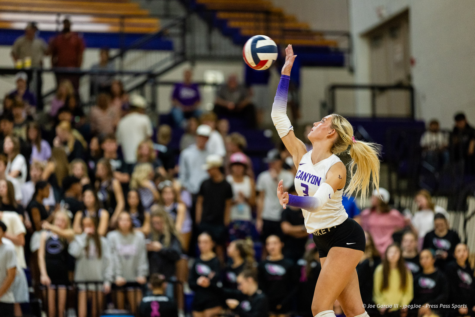 Female volleyball player in white and purple Canyon jersey (number 3) reaches up to contact the ball during a match, with a crowded gym audience in the background.