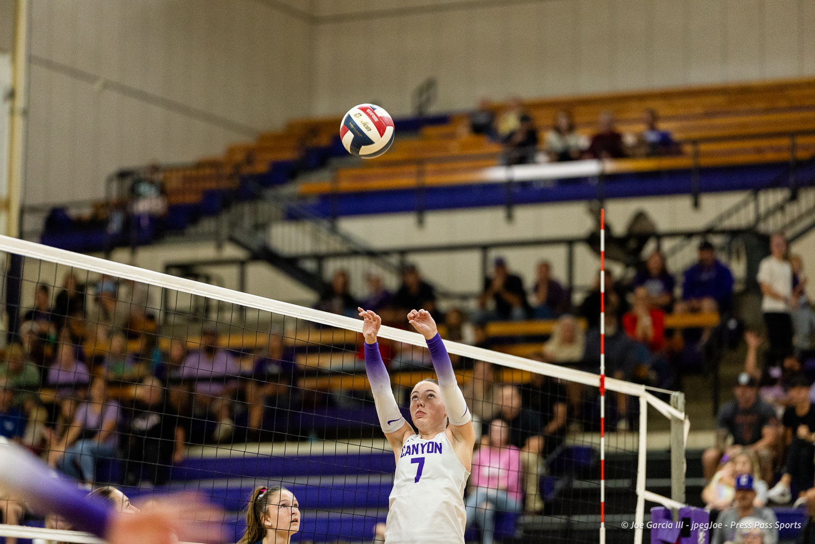 Volleyball player in a white Canyon jersey #7 reaches up to hit the ball over the net as spectators watch from the bleachers.