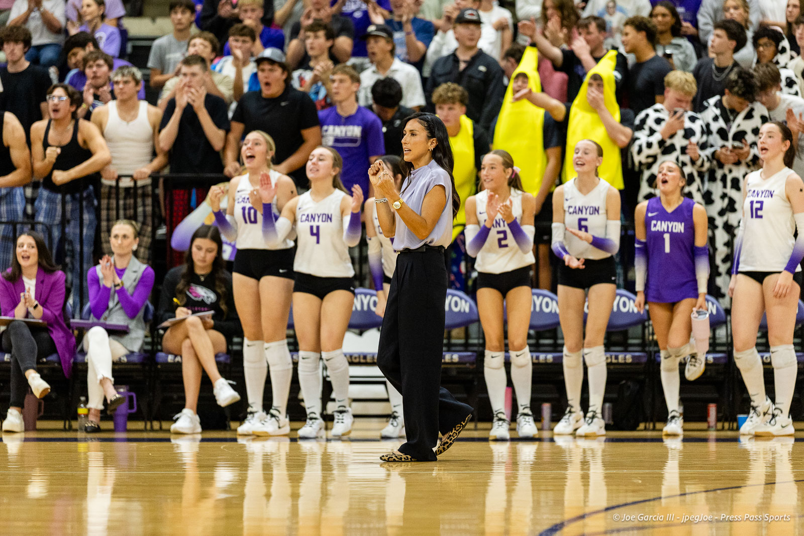 Coach walking across a gym floor while players cheer behind her in Canyon purple and white uniforms.