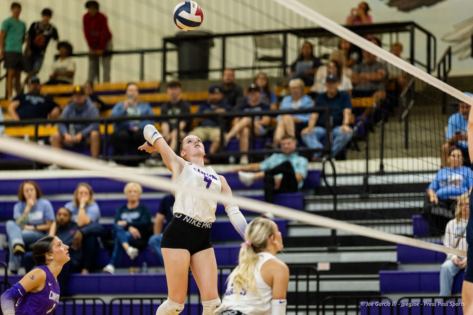 Female volleyball player in a white jersey spikes the ball over the net during a match, with spectators in purple bleachers watching.