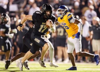 Football player in a black Bisons uniform runs with the ball, pursued by a defender in yellow and blue under stadium lights.