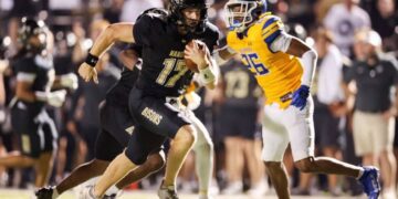 Football player in a black Bisons uniform runs with the ball, pursued by a defender in yellow and blue under stadium lights.