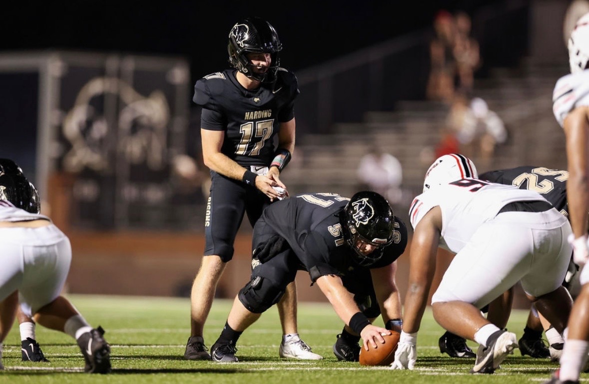 Football play at the line of scrimmage: the center in black snaps the ball to the quarterback under stadium lights.