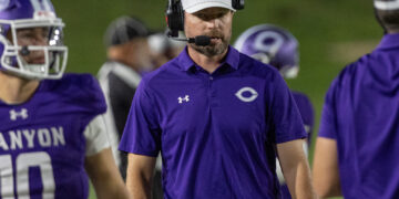 Football coach wearing a purple polo and headset on the sideline during a game at night.