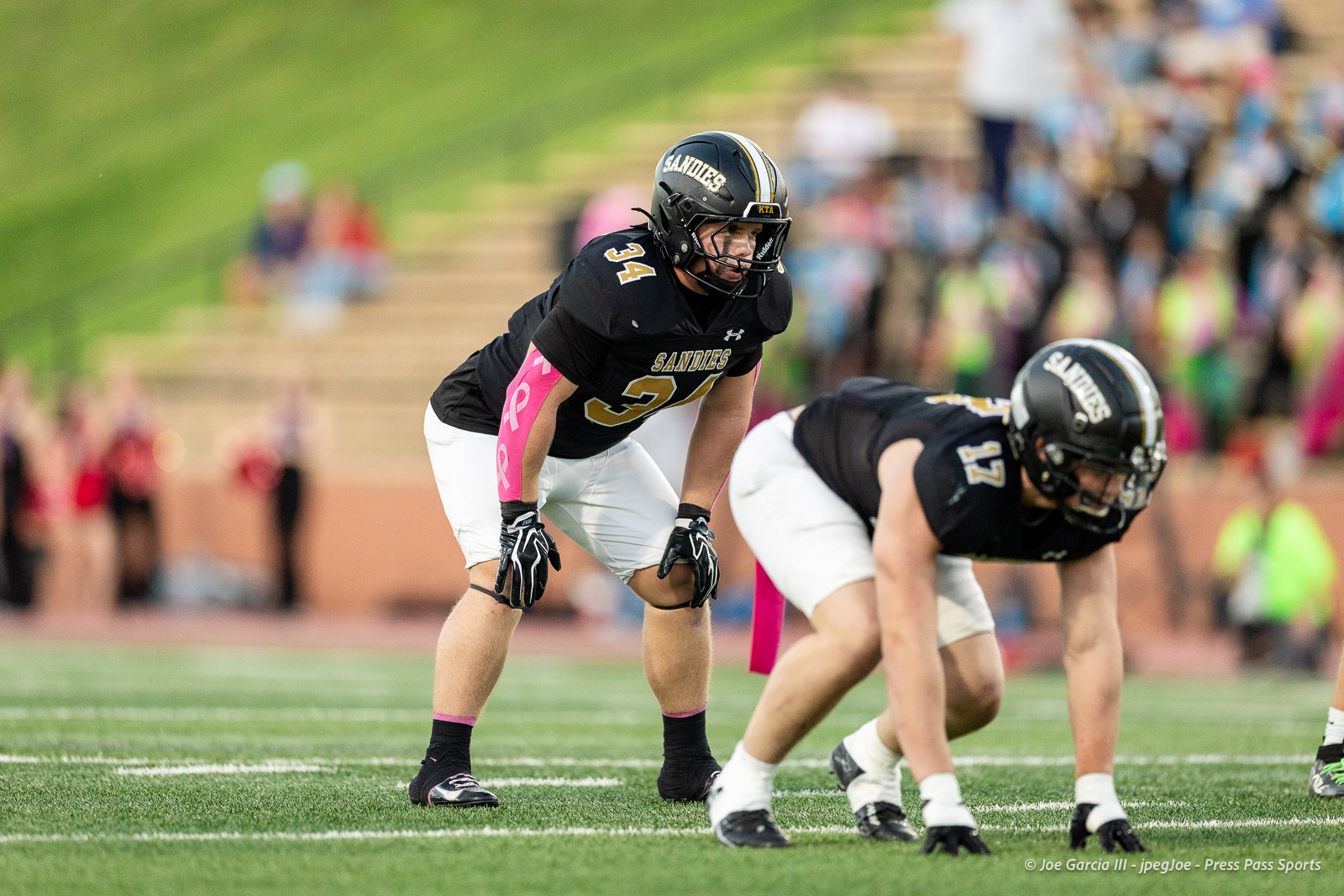 lubbock-monterey-vs-amarillo-high-football-jpegJoe-pps-10 | Press Pass Sports Sandies football players line up on the offensive/defensive line at the snap, black uniforms with pink armbands, focused on the play as the crowd watches in the blurred background