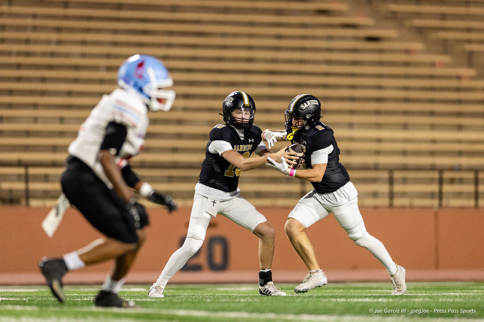 lubbock-monterey-vs-amarillo-high-football-jpegJoe-pps-16 | Press Pass Sports Two Sandies players in black jerseys converge on the football as one hands off to the other on a green field. The opposing player in white rushes in from the left near empty bleachers.