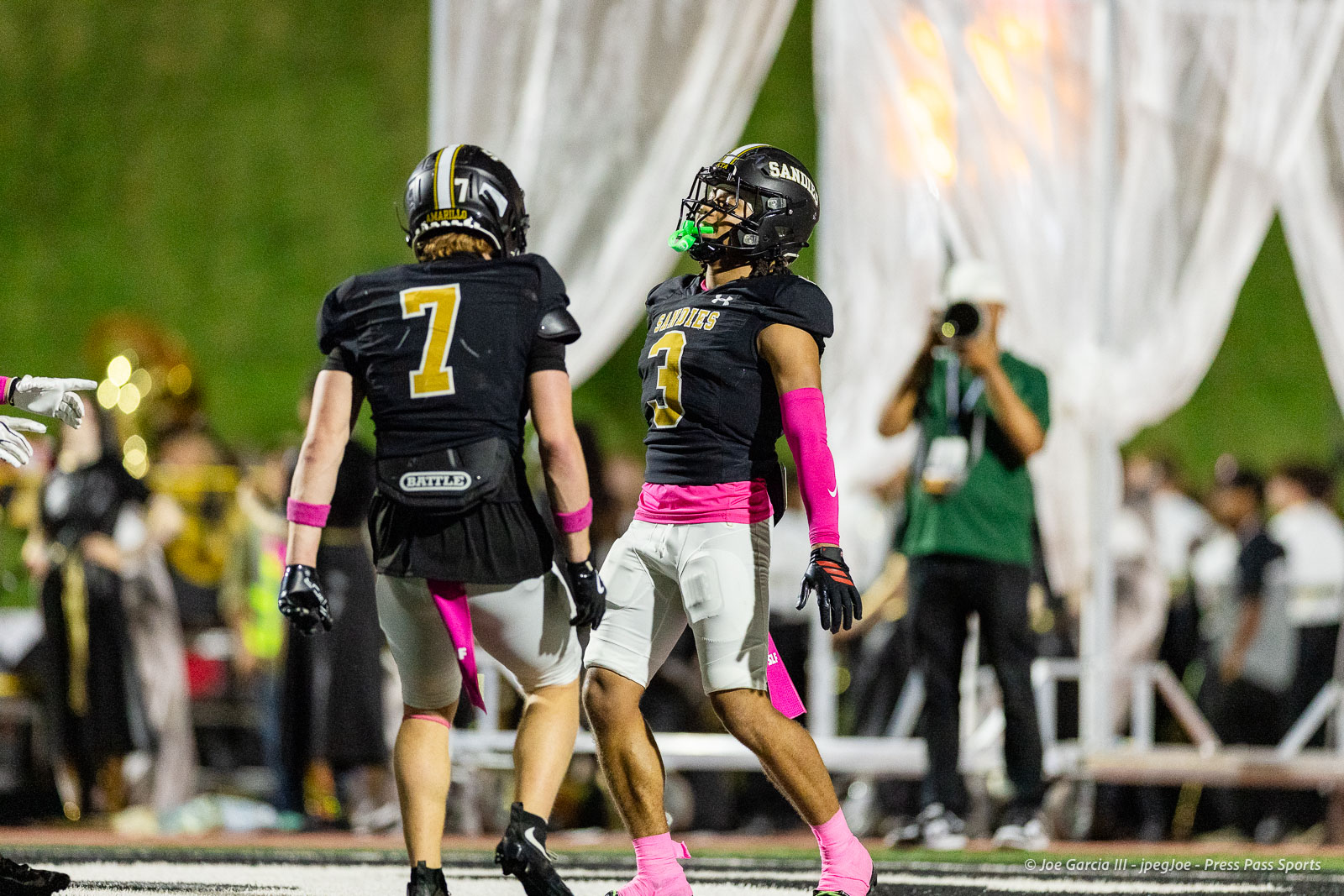 lubbock-monterey-vs-amarillo-high-football-jpegJoe-pps-22 | Press Pass Sports Two football players in black uniforms with yellow numbers (7 and 3), pink accessories, on a lit football field with a photographer in the background.