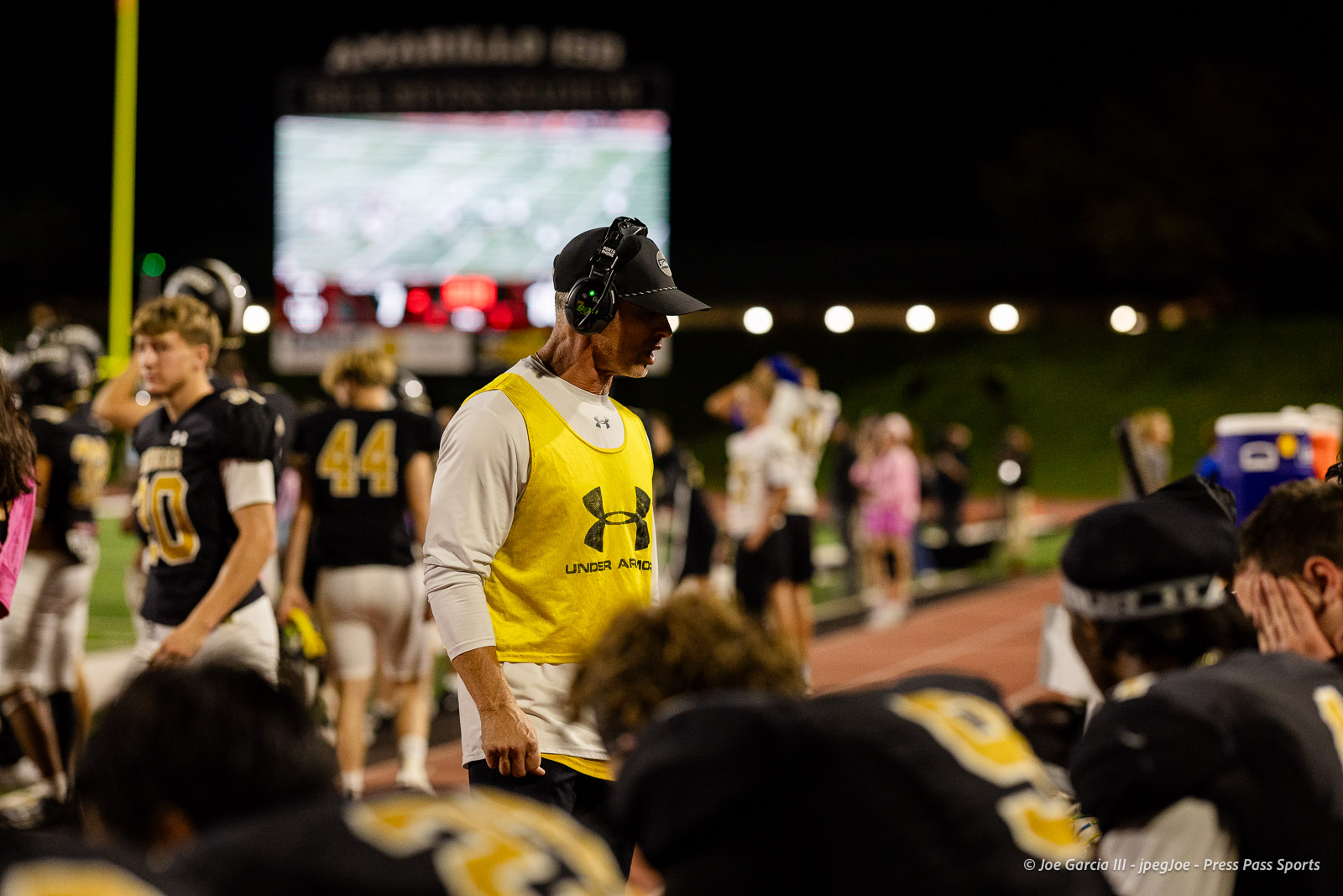 lubbock-monterey-vs-amarillo-high-football-jpegJoe-pps-24 | Press Pass Sports Night football sideline: coach in yellow pinnie with headset, surrounded by players, blurred scoreboard in the background