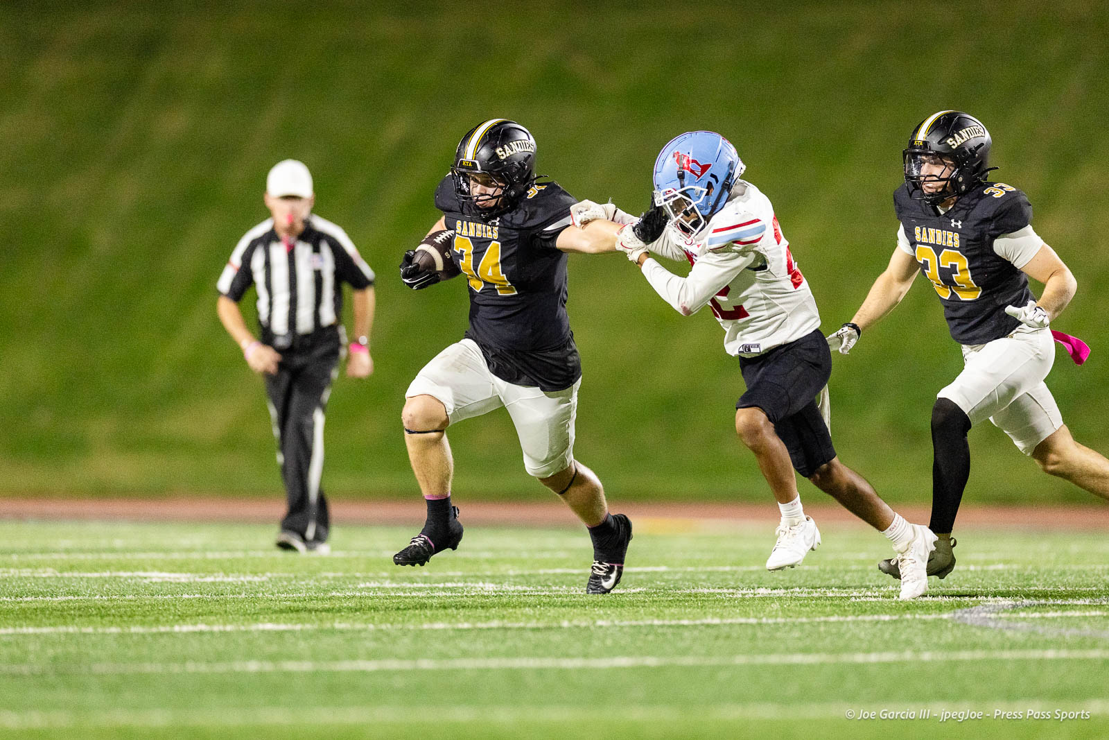 lubbock-monterey-vs-amarillo-high-football-jpegJoe-pps-28 | Press Pass Sports Sandies player (No. 34) carries the football as a defender in a white jersey pursues on a green field during a night game, referee in background.
