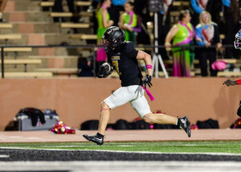 lubbock-monterey-vs-amarillo-high-football-jpegJoe-pps-30 | Press Pass Sports Football player in black jersey #7 running with ball toward end zone; pink armband and ribbon on white shorts on a turf field.