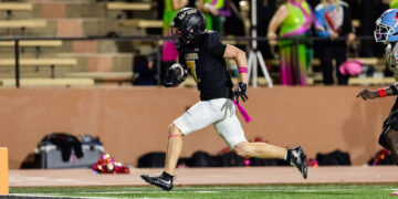 Football player in black jersey #7 running with ball toward end zone; pink armband and ribbon on white shorts on a turf field.