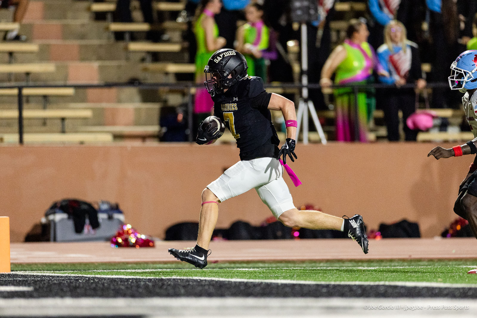 lubbock-monterey-vs-amarillo-high-football-jpegJoe-pps-30 | Press Pass Sports Football player in black jersey #7 running with ball toward end zone; pink armband and ribbon on white shorts on a turf field.